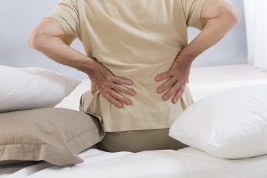 Man sitting in bed with pillows at his right and left side clutching his lower back, used to explain getting an MRI for back pain