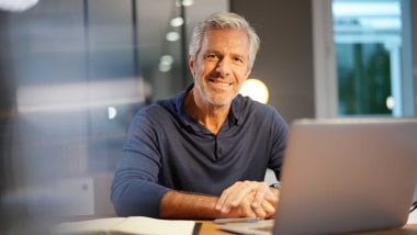 Photograph of a man in a blue shirt smiling at the camera while sitting in front of a laptop, used to describe breast cancer in men