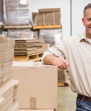 Man standing toward the camera smiling while leaning his arm on a box in an office setting, used to explain varicose veins in men