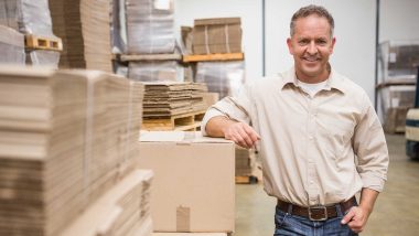 Man standing toward the camera smiling while leaning his arm on a box in an office setting, used to explain varicose veins in men