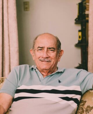 An older man sitting on a floral armchair, smiling and facing the camera, used to explain the signs of Alzheimer's disease