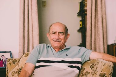 An older man sitting on a floral armchair, smiling and facing the camera, used to explain the impact of Alzheimer's disease