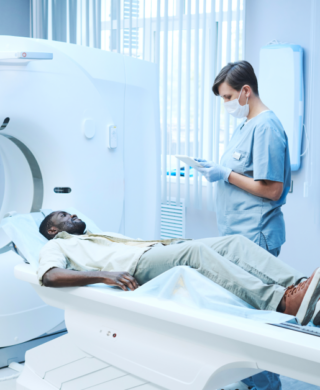 A patient lies on a CT scanner bed while a healthcare professional in scrubs and a mask reviews information on a tablet beside the machine.
