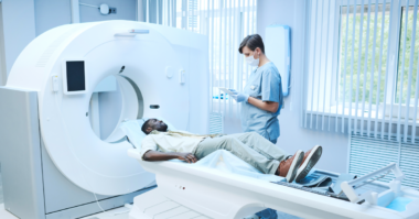 A patient lies on a CT scanner bed while a healthcare professional in scrubs and a mask reviews information on a tablet beside the machine.
